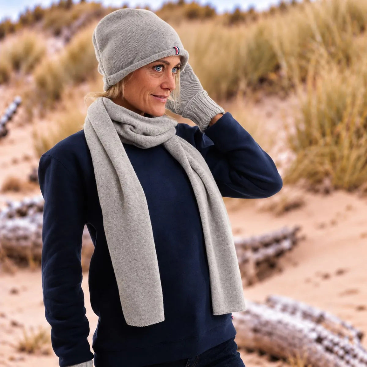 Femme blonde portant un bonnet tombant en cachemire et laine Gris Gavarnie, l'écharpe et les gants assortis, dans les dunes – PALÂBA
