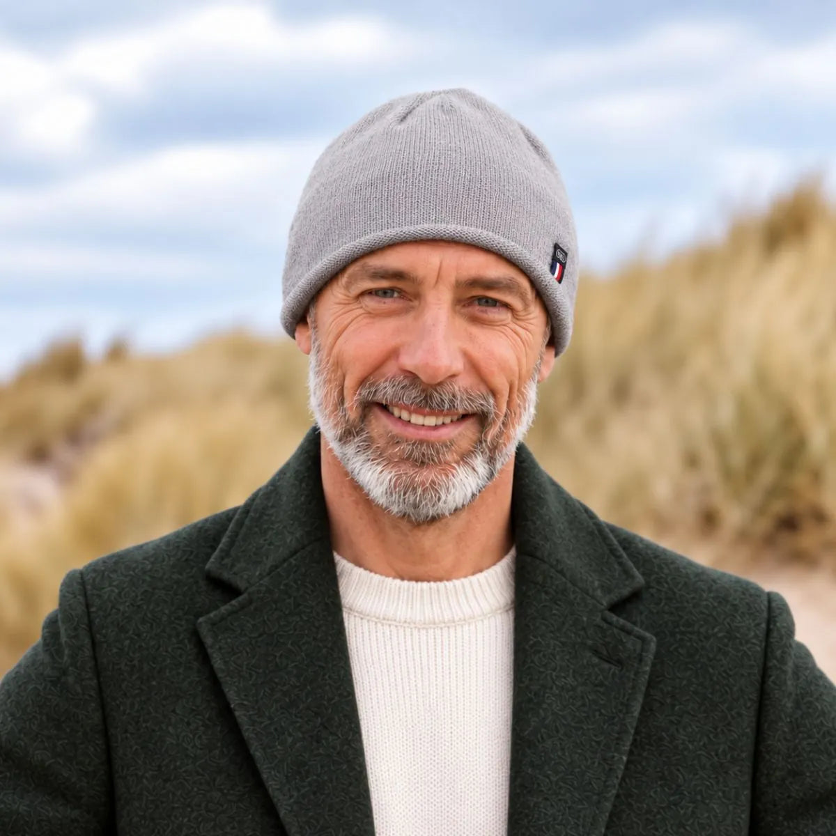 Homme barbu portant un bonnet en cachemire et laine Gris Gavarnie à bord roulotté, manteau vert foncé, dans les dunes – PALÂBA