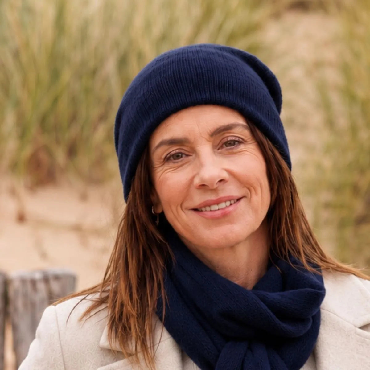 Femme souriante portant un bonnet et une écharpe en cachemire et laine Marine Léman, manteau crème, dans les dunes – PALÂBA