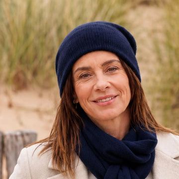 Femme souriante portant un bonnet et une écharpe en cachemire et laine Marine Léman, manteau crème, dans les dunes – PALÂBA