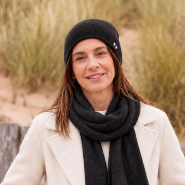 Femme souriante portant un bonnet et une écharpe en cachemire et laine Noir Val d'Isère, manteau blanc, dans les dunes – PALÂBA