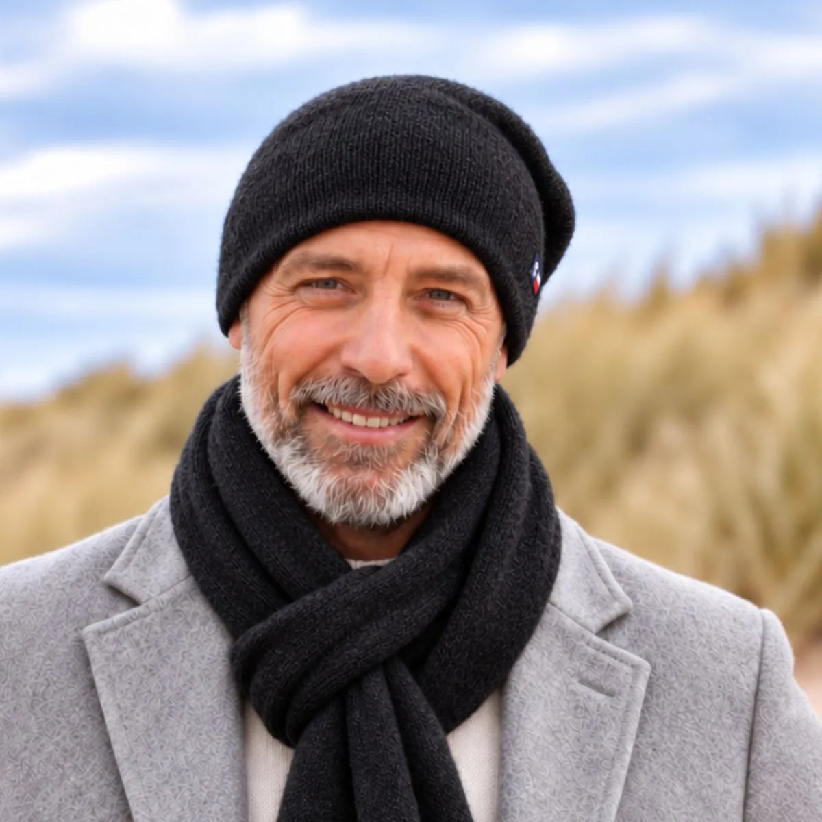 Homme barbu portant un bonnet et une écharpe en cachemire et laine Noir Val d'Isère, manteau gris, dans les dunes – PALÂBA