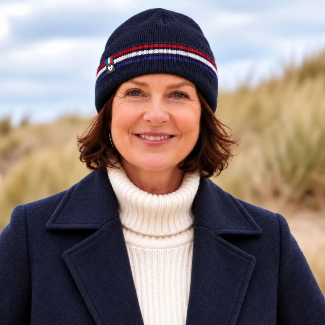 Femme souriante portant un bonnet en laine bleu marine à revers rayé bleu-blanc-rouge, manteau marine, dans les dunes – PALÂBA