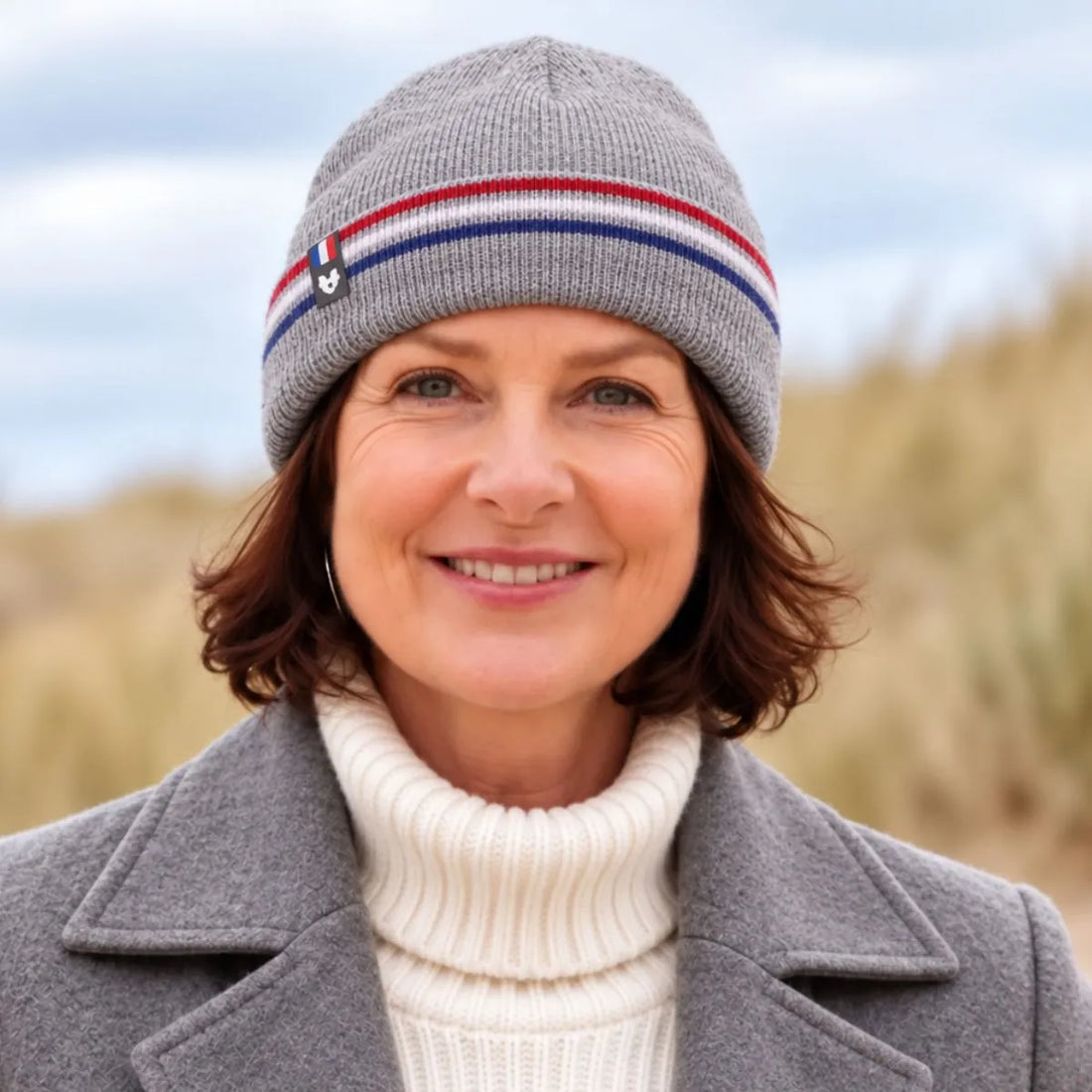 Femme souriante portant un bonnet en laine gris à revers rayé bleu-blanc-rouge, col roulé crème, dans les dunes – PALÂBA