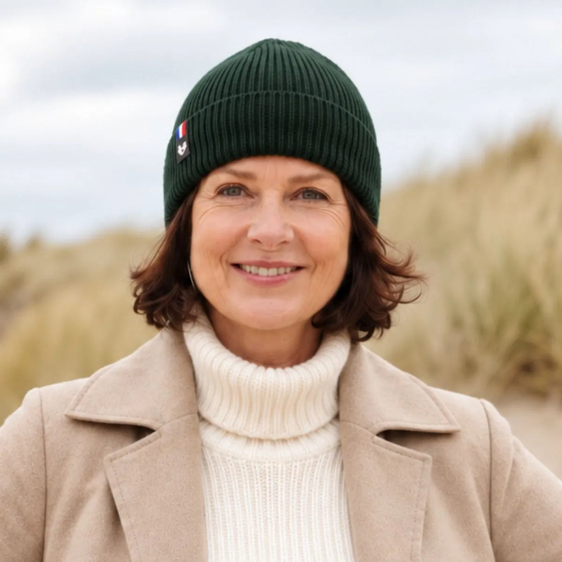 Femme souriante portant un bonnet en laine mérinos vert Forêt Bauges, manteau beige et col roulé crème, dans les dunes – PALÂBA