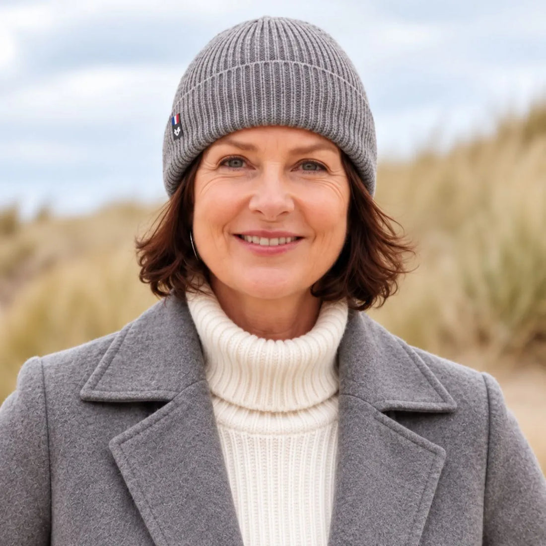 Femme souriante portant un bonnet en laine mérinos Gris Gavarnie à revers, manteau gris et col roulé crème, dans les dunes – PALÂBA