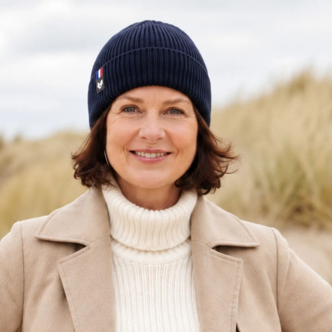 Femme souriante portant un bonnet en laine mérinos Marine Léman, manteau beige et col roulé crème, dans les dunes – PALÂBA