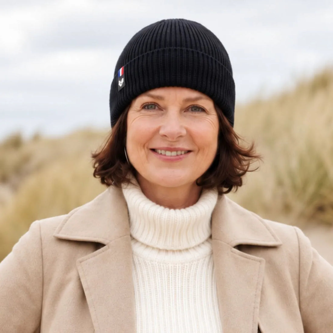 Femme souriante portant un bonnet en laine mérinos Noir Val d'Isère, manteau beige et col roulé crème, dans les dunes – PALÂBA