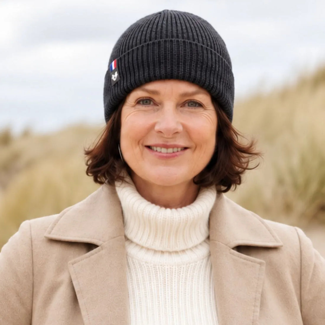 Femme souriante portant un bonnet en laine mérinos Roc Meije, manteau beige et col roulé crème, dans les dunes – PALÂBA