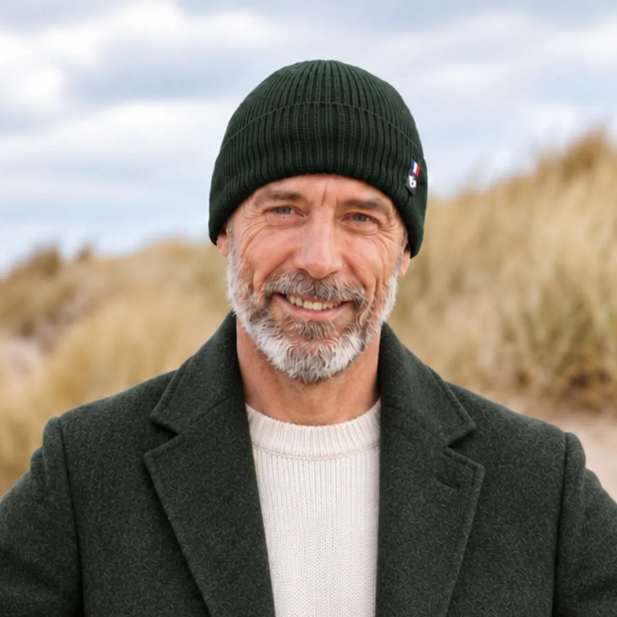 Homme barbu portant un bonnet en laine mérinos vert Forêt Bauges avec un manteau vert foncé, dans les dunes en bord de mer – PALÂBA