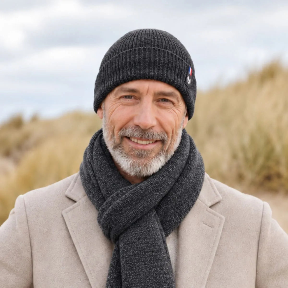Homme barbu portant un bonnet en laine mérinos Roc Meije et une écharpe anthracite assortie, manteau beige, dans les dunes – PALÂBA