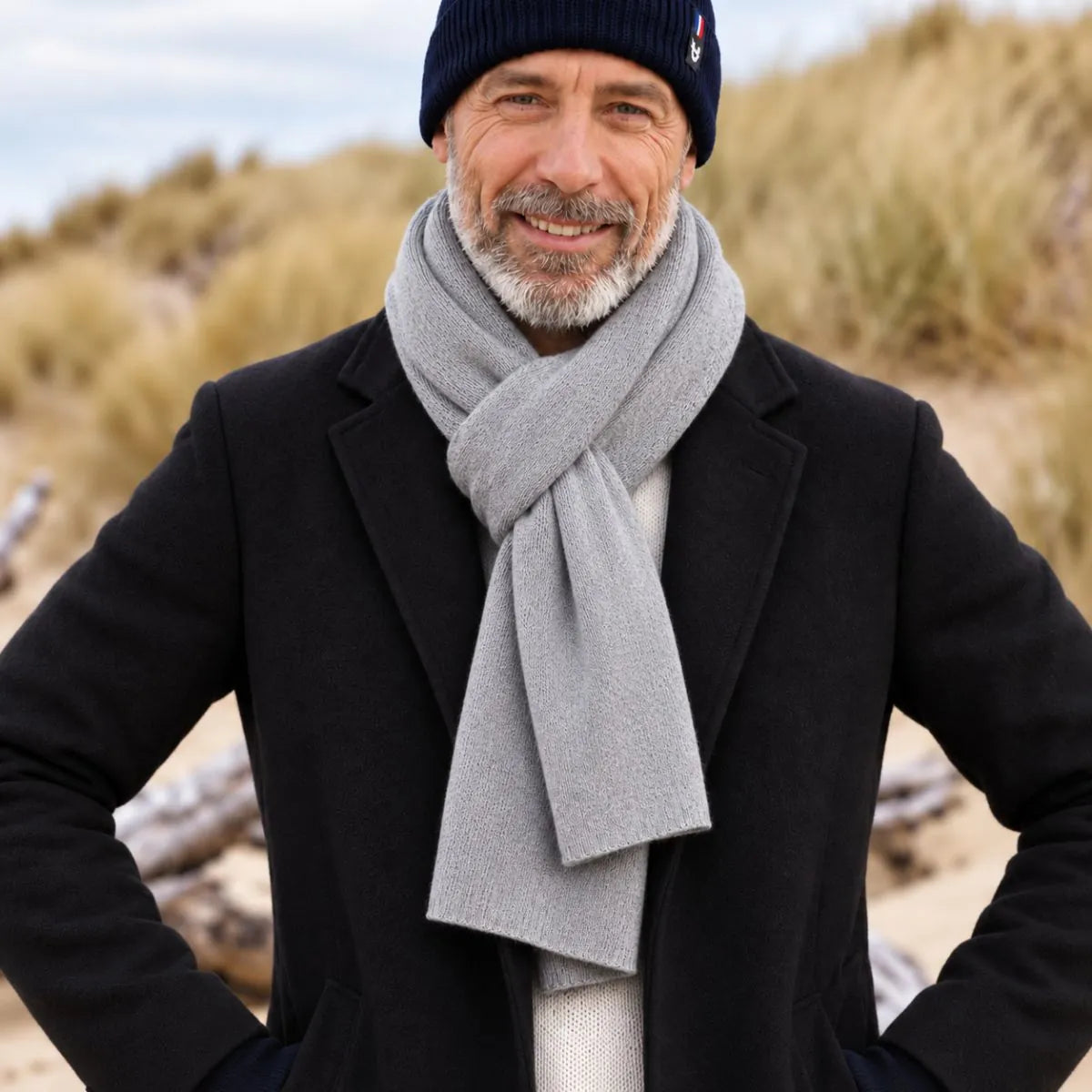 Homme barbu portant une écharpe en cachemire et laine Gris Gavarnie nouée sur un manteau noir, dans les dunes – PALÂBA