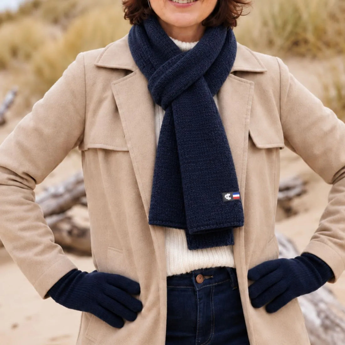 Femme en manteau beige portant une écharpe en laine mérinos Marine Léman nouée et les gants bleu marine assortis, sur la plage – PALÂBA