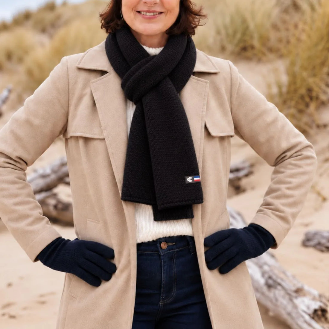 Femme en manteau beige portant une écharpe en laine mérinos Noir Val d'Isère nouée, sur la plage – PALÂBA