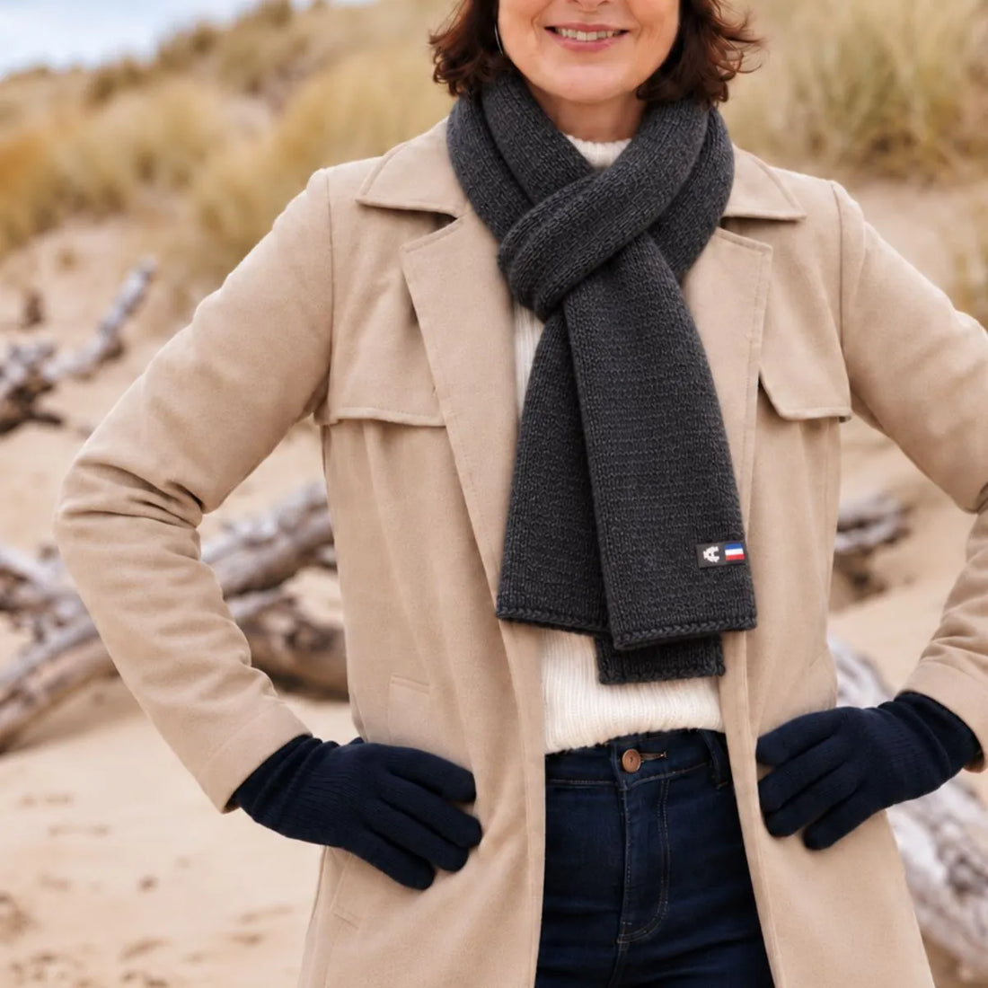 Femme en manteau beige portant une écharpe en laine mérinos Roc Meije nouée, dans les dunes en bord de mer – PALÂBA