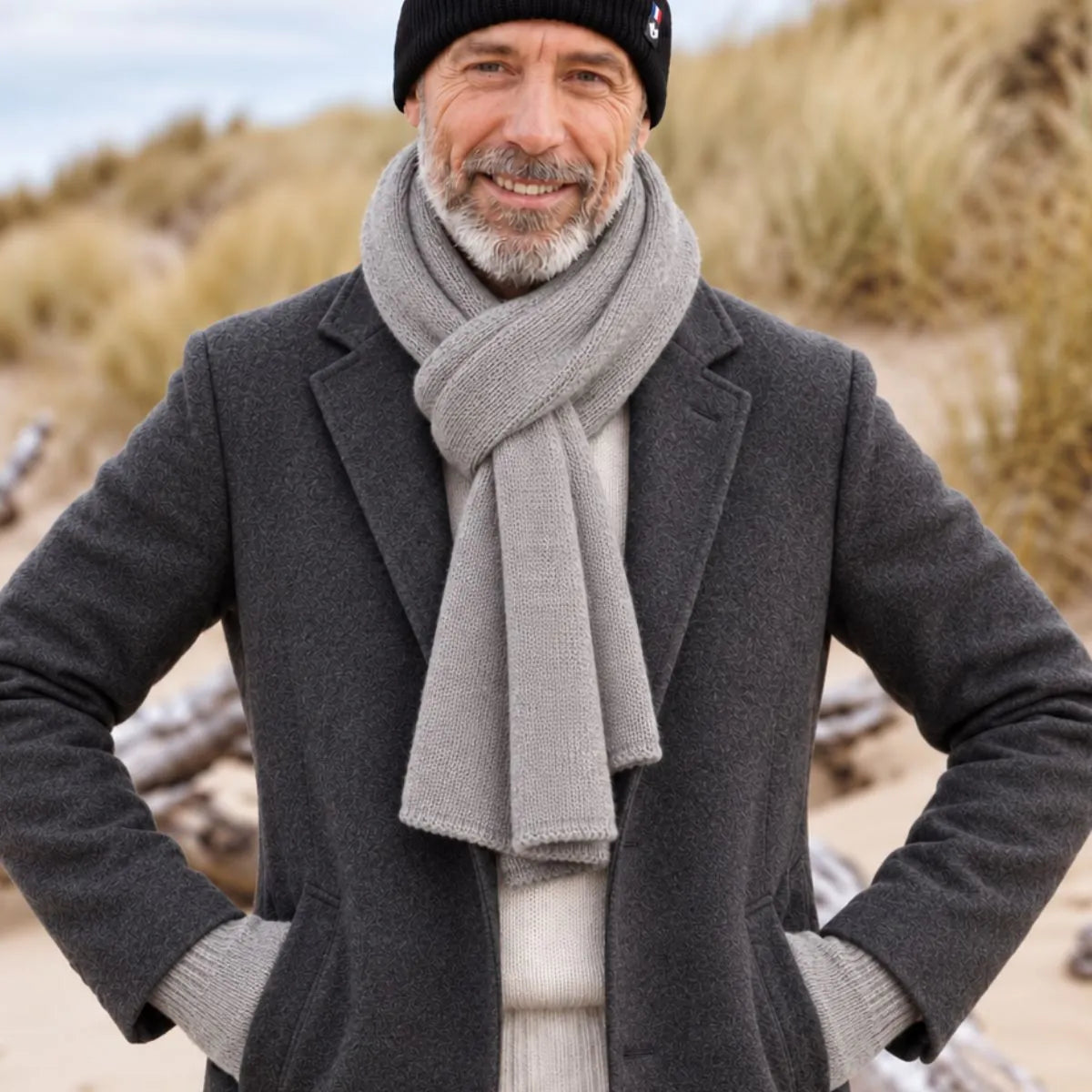 Homme barbu en manteau anthracite portant une écharpe en laine mérinos Gris Gavarnie nouée et un bonnet noir, dans les dunes – PALÂBA