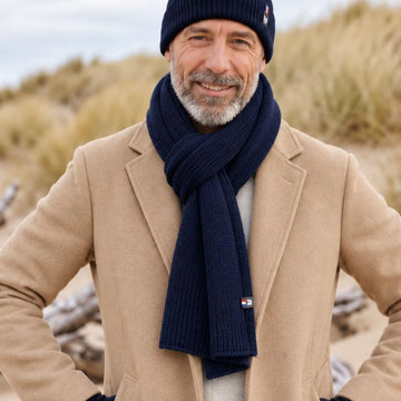 Homme barbu portant une écharpe en laine mérinos Marine Léman et le bonnet bleu marine assorti, manteau camel, dans les dunes – PALÂBA