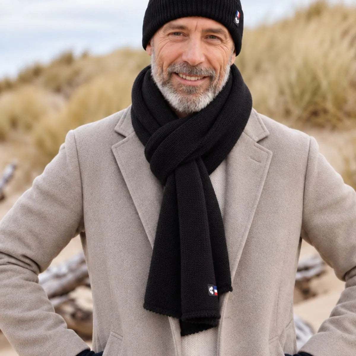 Homme barbu portant une écharpe en laine mérinos Noir Val d'Isère et le bonnet noir assorti, manteau beige, dans les dunes – PALÂBA