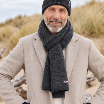 Homme barbu portant une écharpe en laine mérinos Roc Meije nouée sur un manteau beige, bonnet assorti, dans les dunes – PALÂBA