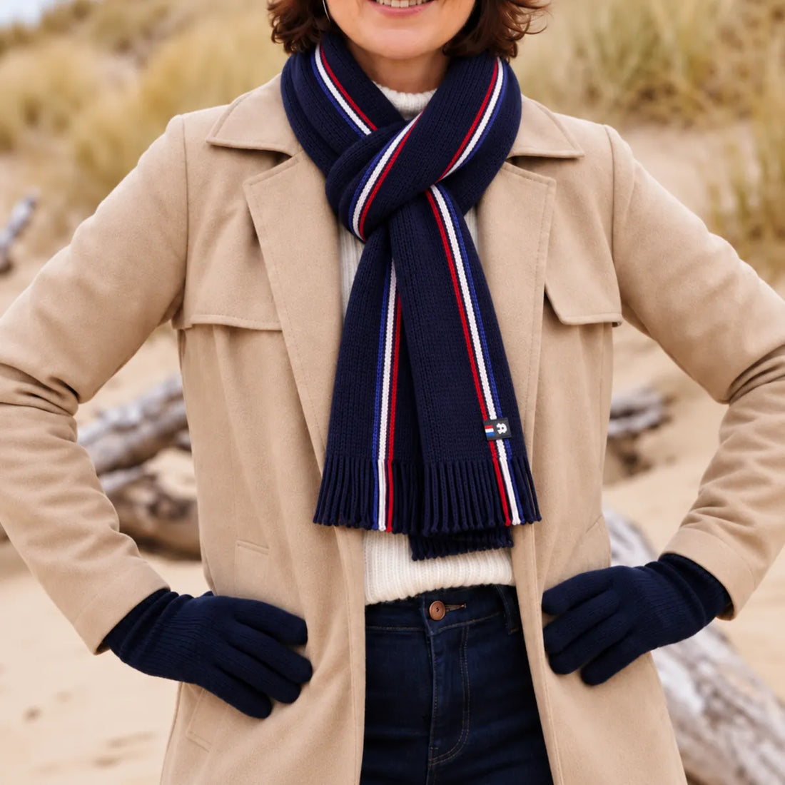 Femme en trench beige portant une écharpe tricolore bleu marine à franges et des gants marine, sur la plage – PALÂBA
