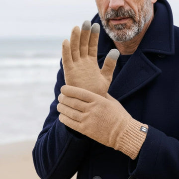 Homme portant des gants tactiles en laine mérinos Camel Aubrac avec un manteau bleu marine, face à la mer – PALÂBA