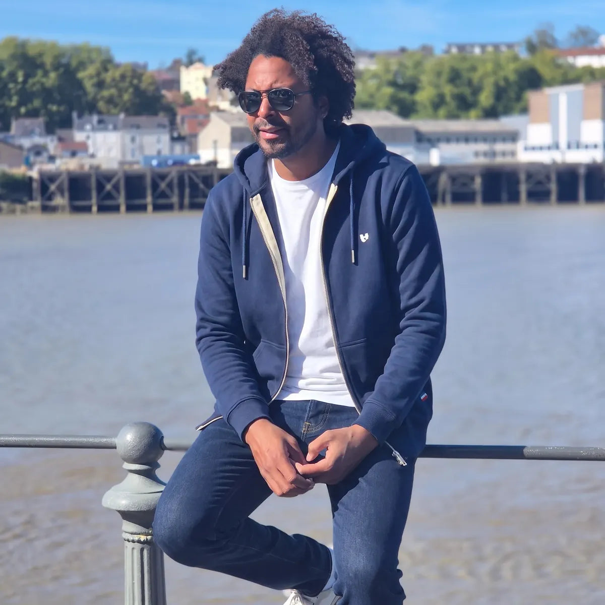 Homme portant le hoodie zippé molletonné marine ouvert sur un t-shirt blanc et un jean brut, au bord de l'eau – PALÂBA