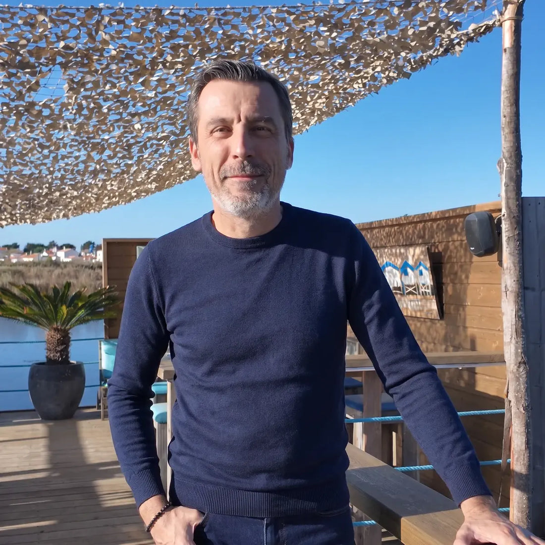 Homme portant le pull bleu marine en laine mérinos et un jean brut sur une terrasse au soleil – PALÂBA