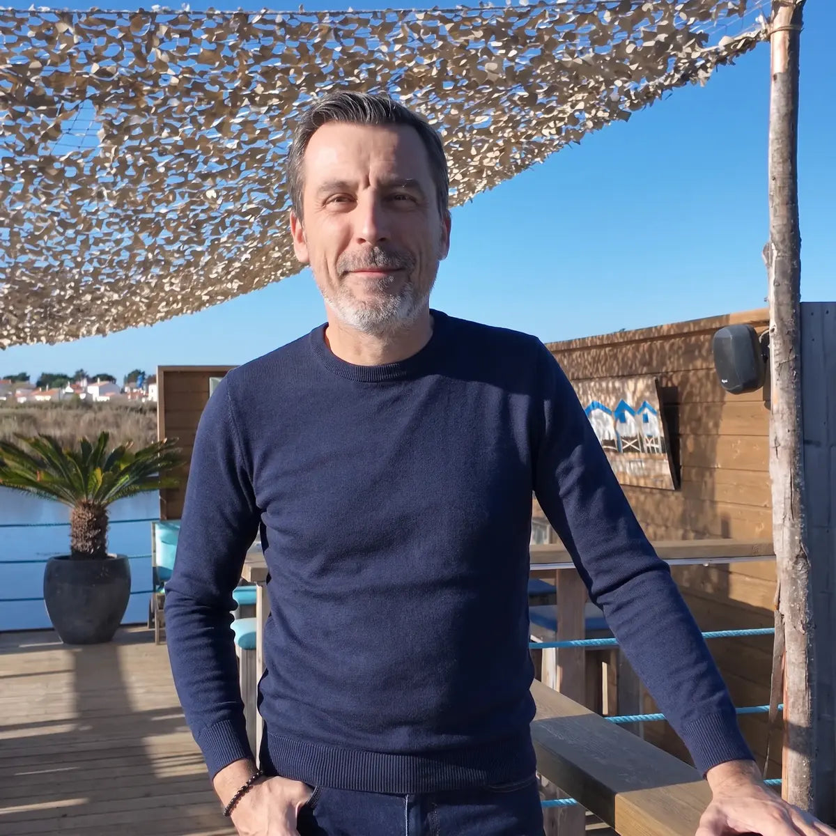 Homme portant le pull bleu marine en laine mérinos et un jean brut sur une terrasse au soleil – PALÂBA