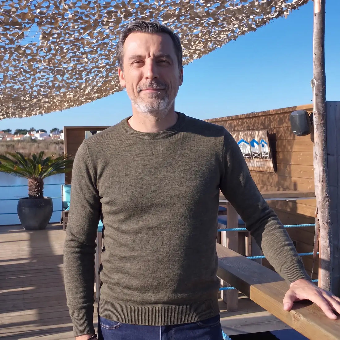 Homme souriant portant le pull kaki en laine mérinos sur une terrasse en bois face à l'eau – PALÂBA