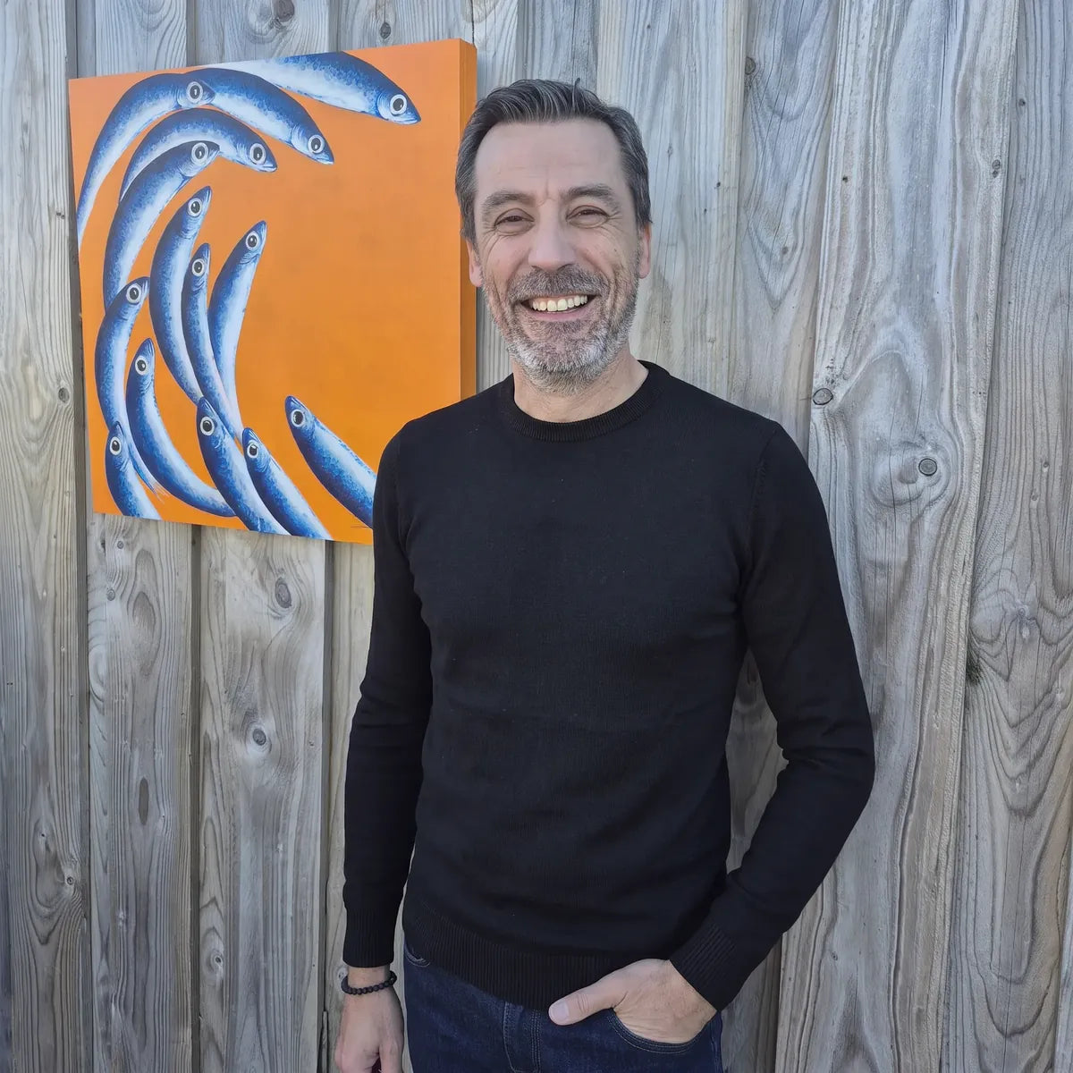 Homme souriant en pull noir en laine mérinos et jean brut devant un tableau de poissons – PALÂBA