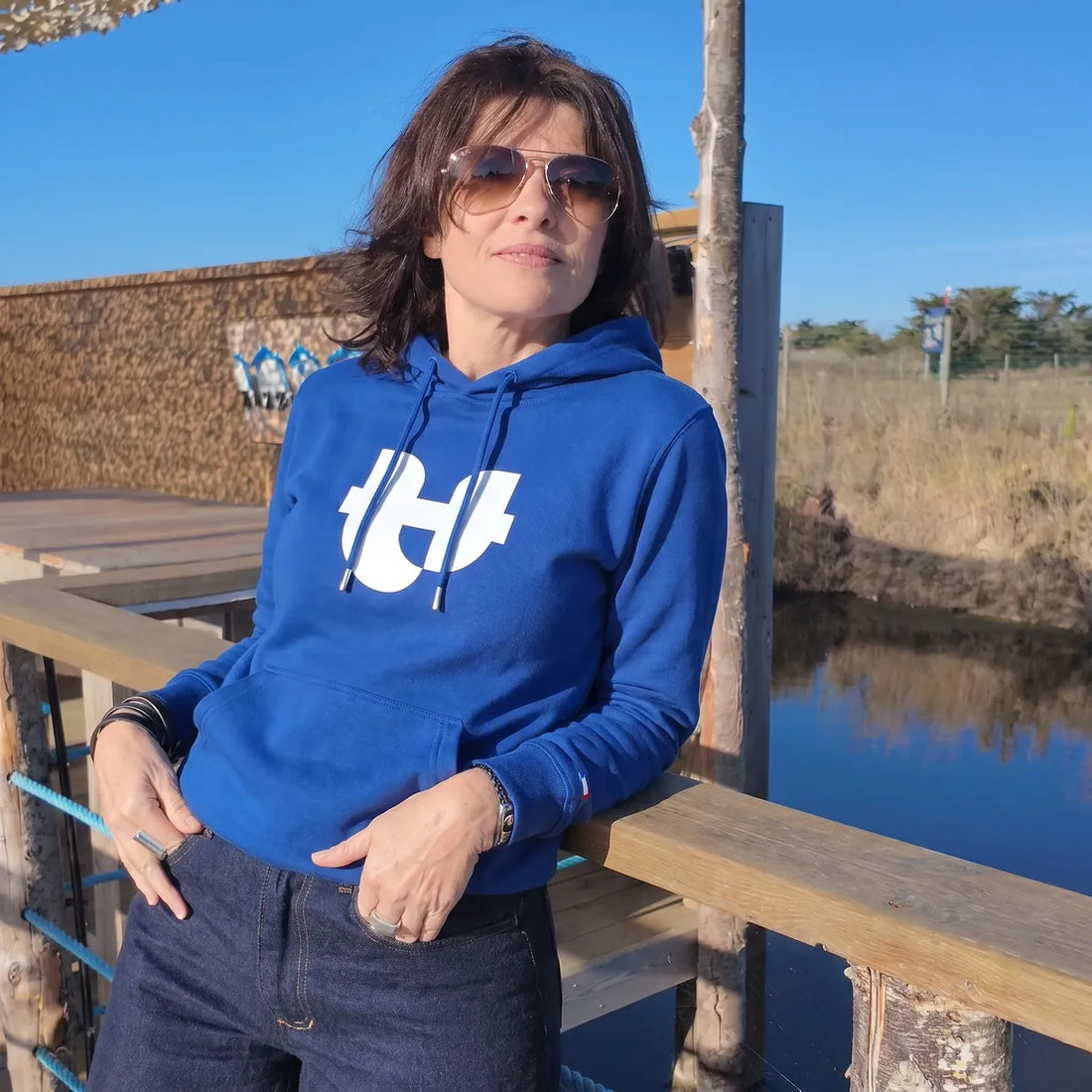 Femme portant le sweat à capuche bleu électrique au grand logo PALÂBA blanc avec un jean brut, sur un ponton au bord de l'eau – PALÂBA