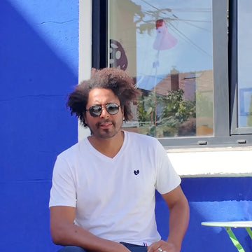 Homme portant le t-shirt col V blanc en coton bio et des lunettes de soleil, devant un mur bleu – PALÂBA